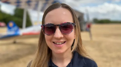 A woman wearing sunglasses is standing in front of a small plane on a field.