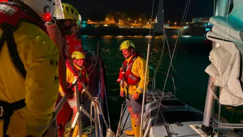 RNLI crew members in safety gear board a sailboat at a harbour at night, illuminated by deck and dock lights.