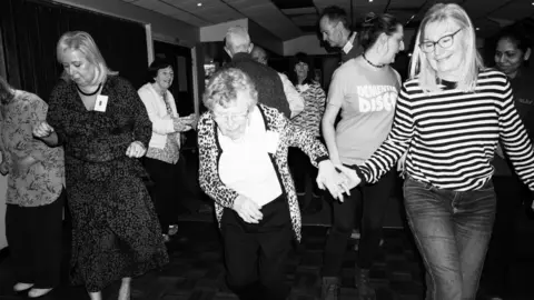Fern Deacon/Stare Society Studio Some older people are dancing and holding hands with younger people in this black and white photo. Three of those shown in this photo are smiling. One older woman is concentrating while dancing.