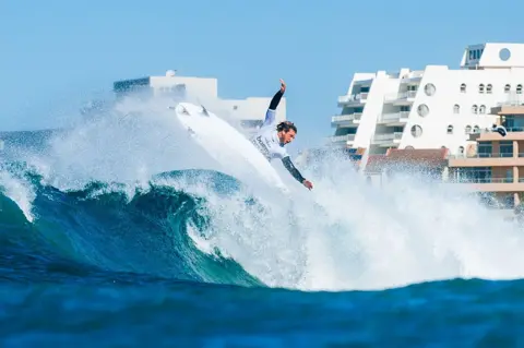 KODY MCGREGOR / WORLD SURF LEAGUE / GETTY IMAGES Ryan Huckabee of the United States crests a wave. Behind him apartment blocks are visible.