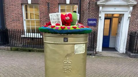 Knitted representations of a red devil, Champagne bottle, a heart shape and a Valentines card. They are sat on top of a gold post box. There is a brick building in the background of the photo.