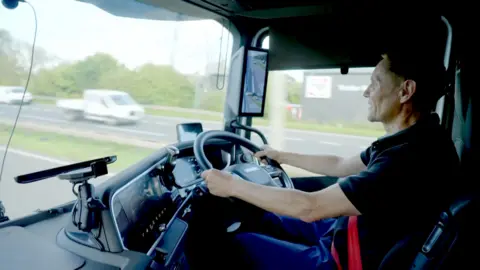 A lorry driver pictured inside his cab, sitting at the steering wheel, as he drives along a dual carriageway, with the view outside of passing vehicles slightly blurred as his lorry moves along