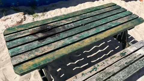 A picture of a picnic bench that is in a state of disrepair. There is a large burnt scorch mark on the table from a barbecue. 