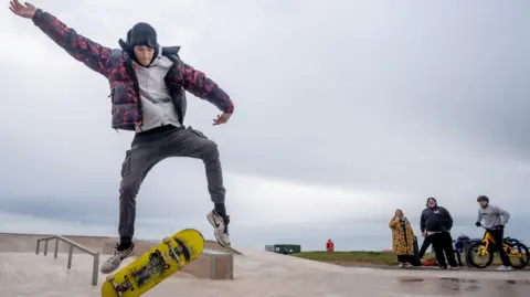 Tom Kay A young man doing a trick with a skateboard at the new Maryport skatepark. There are three bystanders watching, including a man on a BMX. 