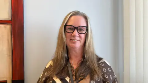 Melanie Guest is sitting in an office, with a pale wall behind her, a piece of art to her left and a window blind to the right. She's wearing a patterned shirt and black-framed glasses.
