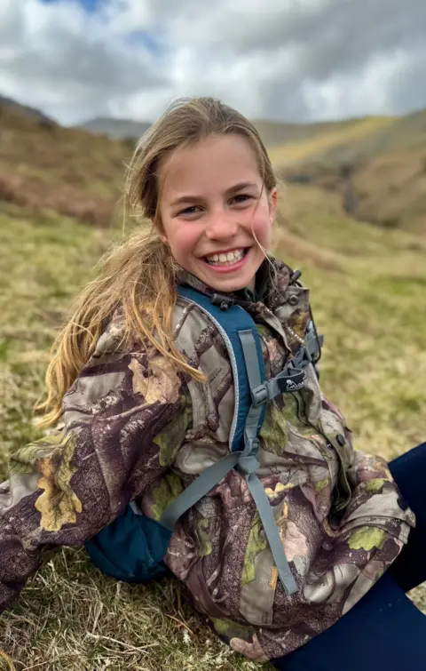 Kensington Palace Princess Charlotte smiles to the camera while sitting on a hillside wearing a camoflage jacket and blue backpack