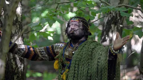 A man in a patterned jumper with a green shawl draped over his shoulder wearing some kind of beaded headdress stands with his arms spread wide in a woodland