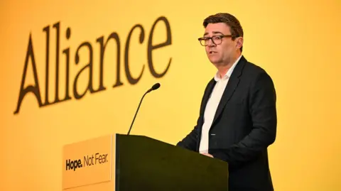 PA Media Andy Burnham at Alliance conference standing at a yellow podium saying 'hope not fear'. Behind him is a yellow background with 'Alliance' written in black. Andy Burnham has short dark hair and glasses. He is wearing a black suit with a white shirt.