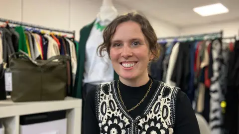 Imy smiles into the camera, wearing a black long-sleeve top and a white and black waistcoat with a gold chain. She has curly, brown hair tied up. Ranks of clothing are blurred in the image behind her. 