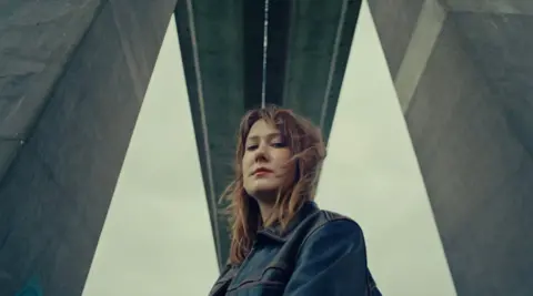 Halo Red-haired Ellena Russell, wearing a blue denim jacket, looks down to the camera, from underneath the Orwell Bridge in Ipswich. The concrete pillars she is between are grey. 