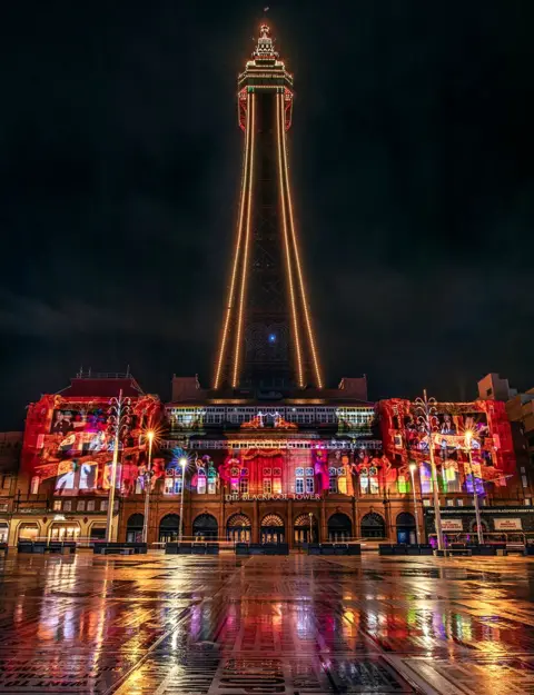 VisitBlackpool Blackpool Tower, pictured at night lit up with gold bulbs, with additional projections in red and gold on the building at its base.