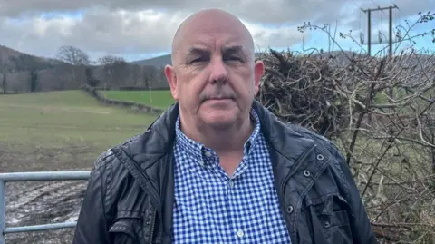 A bald man, wearing a white and blue check shirt and black jacket. He is standing in front of a field. 