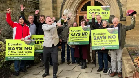 Parish Clerk Adam Shanley cracks open the champagne alongside local residents of Dunstan. He is wearing a beige suit jacket and black trousers.  Residents are holding green signs reading Dunstan Says No to New Build Holiday Accommodation.