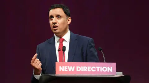 Anas Sarwar speaking at the Labour party conference in Glasgow - he is standing behind a lectern with a sign which has NEW DIRECTION written on it in white letters on a red backdrop. He is wearing a dark suit with a red tie.