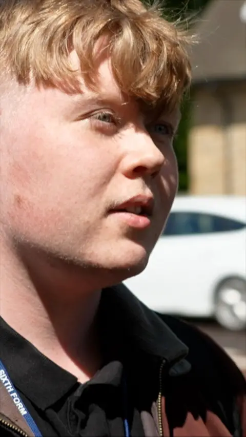 A young man with sandy hair looking off to the camera. There is a brick building behind him.