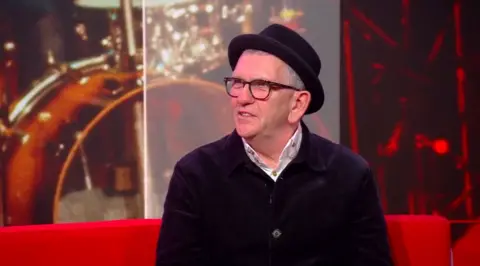 BBC Mike Joyce sits on a red sofa in the BBC North West Tonight studio. He wears a black hat, black glasses and a white shirt under a black jumper