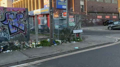 The corner of a road with a street sign that states "Trent Street". It is in front of a wired fence with a graffitied board in the left-hand corner. There is rubbish on the grass next to it. There is a large building in the background and parked cars.