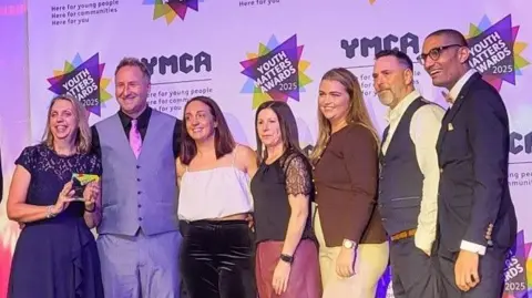 Seven people on a stage smiling at a camera off to the left of this image.