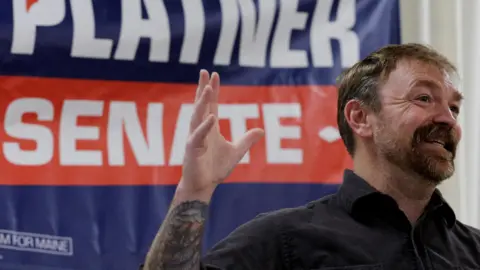 Graham Platner holds a tattoo'd arm up in front of a blue and orange sign reading Platner Senate