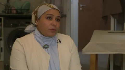 Fozia Malik sat in the living room of her apartment, wearing a cream jacket and light blue headscarf. 