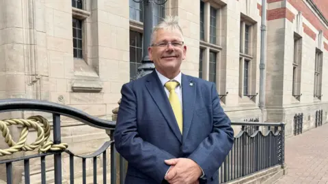 Patrick Allen is standing in front of a light-coloured stone building next to some black railings. He is wearing a blue suit and yellow tie with a white shirt, and smiling at the camera.