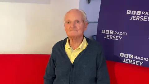 An old man with grey hair and a bald spot on top, wearing a yellow shirt and blue cardigan.