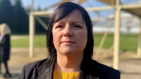 A woman with black hair and a black jacket and yellow top outdoors head and shoulders shot with blurry background