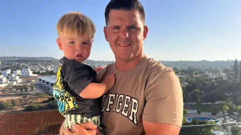 Victoria Thomas Teddie and his father, Ryan, pictured on holiday. Ryan is holding Teddie, Ryan is smiling at the camera and has a tanned t-shirt on with the words "Hilfiger" in block white writing across the front. He has dark cropped hair. His son has a charcoal t-shirt on with a colourful pattern on the back with striped green and white shorts. He has blonde hair. 