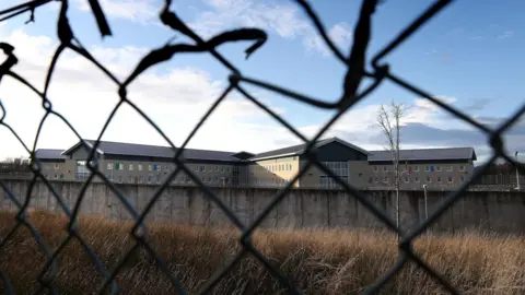PA Media Low Moss Prison buildings behind a high wall, pictured through the wire of fence.