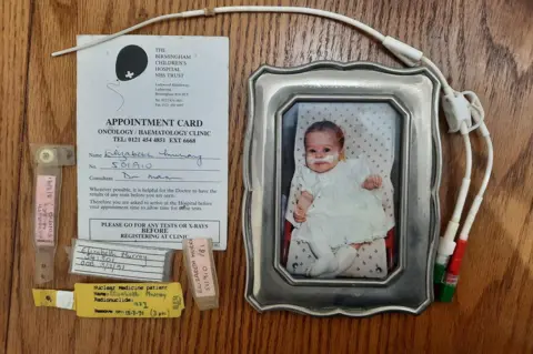 Elizabeth Rees-Gilbert A framed image of Elizabeth as a baby is surrounded by hospital paraphernalia. Her appointment card for Birmingham Children's Hospital is there along with hospital patient wrist bands. A nasal tube can also be seen. 