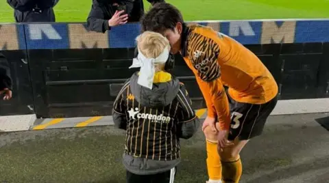 Submitted A football player in a black and orange kit chats to a little boy in a striped shirt and coat with a bandage around his head