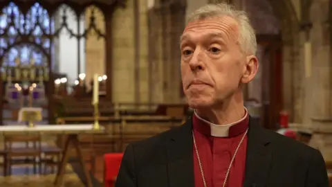 Andrew John in a red shirt and black blazer with a dog collar, looking at the camera in front of an altar. He has grey hair and brown eyes