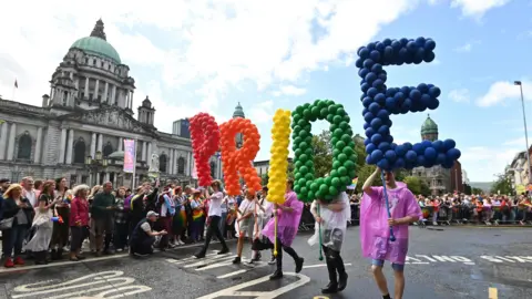 People taking part in a parade hold balloons in the shape of letters spelling out pride. They're in red, orange, yellow, green and blue. People are watching from the street. Belfast City Hall is in the background. 