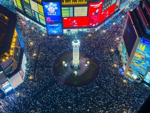 Cheng Xin/Getty Images Pemandangan Monumen Jiefangbei dari udara dikelilingi oleh ribuan orang yang bersuka ria dan papan reklame yang menyala selama perayaan Malam Tahun Baru pada 31 Desember 2024, di Chongqing, Tiongkok. Warga yang bersuka ria datang dalam jumlah besar untuk merayakan tahun baru di Chongqing. 