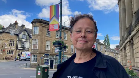 A woman with short, curly hair standing in a street with a rainbow-coloured flag attached to a lamppost in the background (taken 30 June)