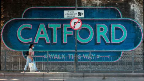 A woman walks in front of a large mural on a wall which says "CATFORD, WALK THIS WAY".