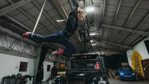 Rave New World A woman is dangling by ropes from an aerial theatre rig attached to the roof of a black taxi