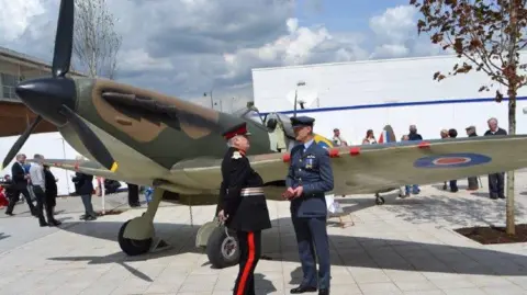 A historic military aircraft, painted in camouflage colors, stands outdoors on a paved area. Several people stand around the plane, including two individuals in formal military-style uniforms engaged in conversation near the wing.