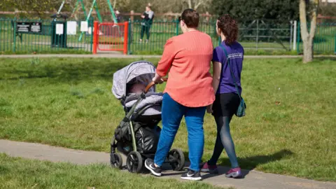 Mothers for Mothers Photo of a mum and a Mothers for mothers volunteer walking with a baby in a pram at a children's park. The mother is pushing the buggy. She is wearing a red blouse with jeans and trainers. Her child is an asleep in the pram. The volunteer is wearing a purple t-shirt and a skirt with tights. They are both walking away from camera with the their backs turned. 