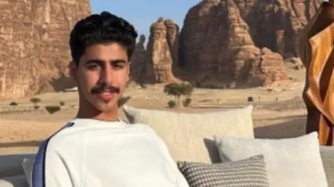Naif Alqassim Mohammed Algasim wearing a white T-shirt with a blue stripe down its shoulders. He is sitting on a sofa with cream pillows next to him. Behind him appears to be a desert scene, with a sandy/rocky surface and tall rock structures.