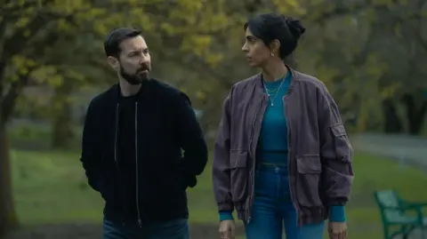 Amazon MGM Studios An image from TV show Fear with Martin Compston and Anjli Mohindra's characters in conversation while walking through a park - he is in a blue hoodie/jacket and she has a purple jacket, aqua top and blue jeans on