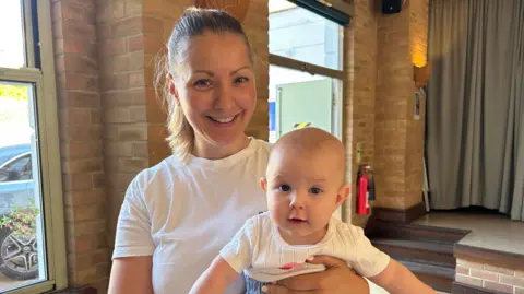 Woman with blond hair in a ponytail, wearing a white t-shirt and holding a baby with a white top, the woman is smiling at the camera and the baby is looking directly at the camera. In the background is a stage with curtains pulled across it.