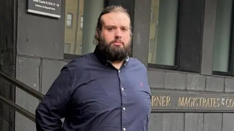 Man stands outside Magistrates court in black shirt with dark beard and hair.