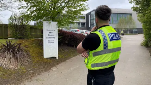 BBC A police officer standing outside Birley Academy with his face turned towards the school building at the end of the drive.