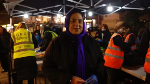 Kian Boyle/BBC A woman stands under a lit outdoor canopy at night, facing the camera. She wears a dark coat, a purple headscarf, and blue gloves, and smiles calmly. Behind her, volunteers in high-visibility vests work at tables. 