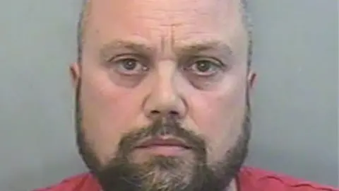 A police mugshot of Craig Short who has a brown beard and is wearing a red top