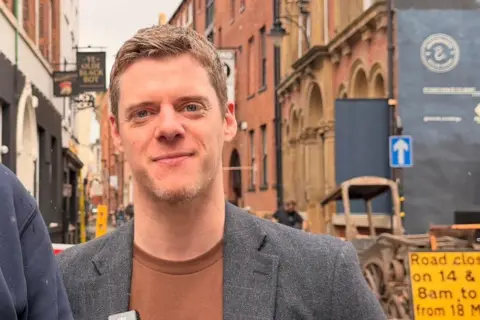 A man standing in a narrow street with high buildings to either side. He is wearing a maroon shirt and grey blazer. He has strawberry blonde hair and a short beard. A yellow "road closure" sign can be seen to the right.