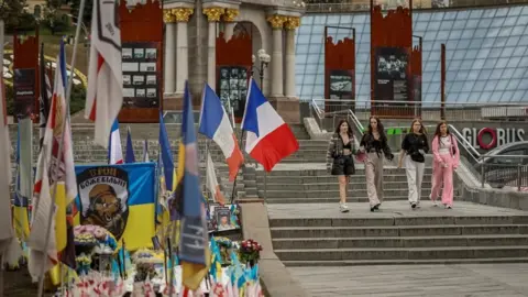 Reuters cilvēki paiet pa maiņas memoriālu kritušajiem Ukrainas aizstāvjiem Kijevas Neatkarības laukumā. Kopā ar ziediem un Ukrainas karogu ir arī citu valstu karogi, ieskaitot Franciju.
