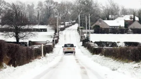 A white car makes its way up a narrow country road in County Antrim, which has a light dusting of snow. Two hedges can be seen on either side of the road and a house is visible in the background.