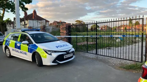 Police car outside gate leading to field - blue and white police tape is across the iron gate
